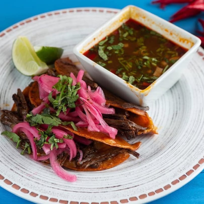 Birria tacos with pickled red onions, cilantro, lime wedges, and a side of consommé.