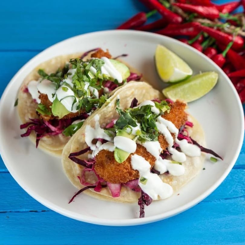 Two fish tacos with red cabbage, avocado, cilantro, and crema, served with lime wedges.