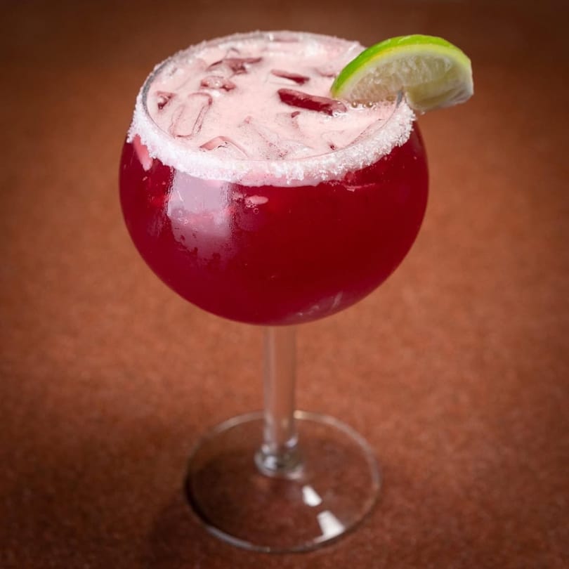 A vibrant red cocktail with ice, a foamy top, and a lime wedge, in a stemmed glass.