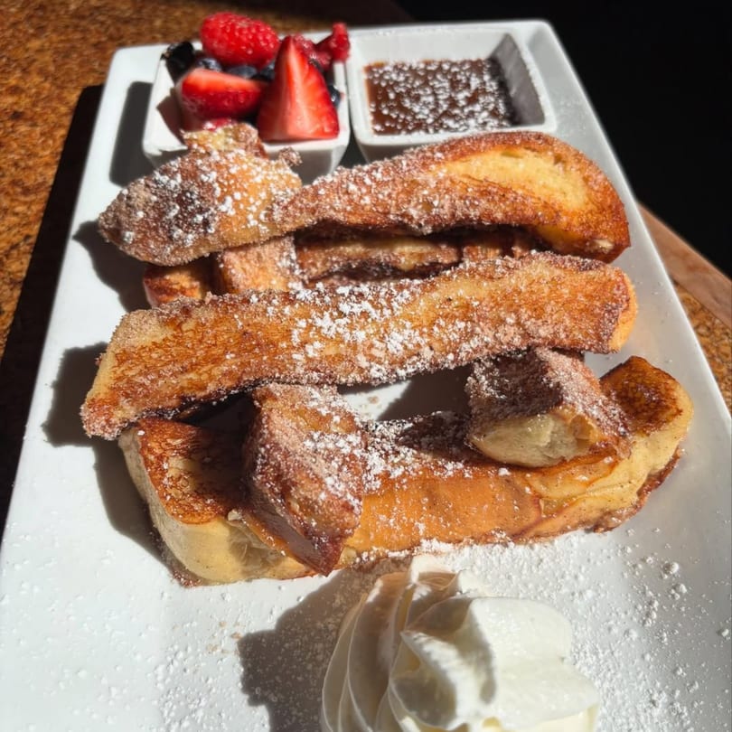 Golden French toast sticks dusted with powdered sugar, served with berries, syrup, and whipped cream.