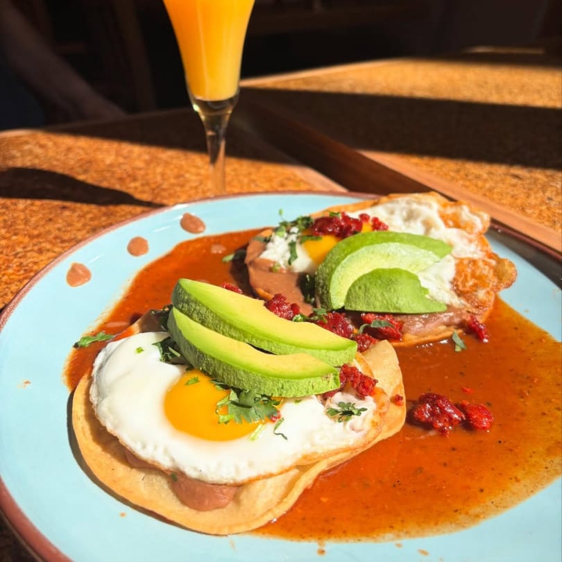 Huevos rancheros with avocado slices and a mimosa on a sunny table.
