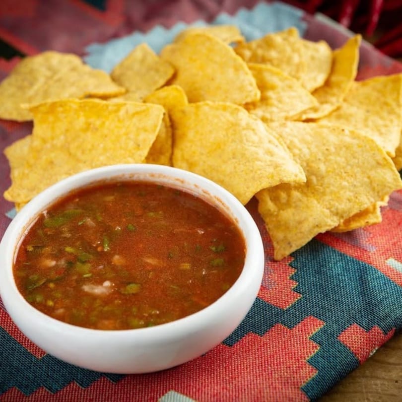 A white bowl of red salsa with green herbs, surrounded by a pile of tortilla chips on a patterned cloth.