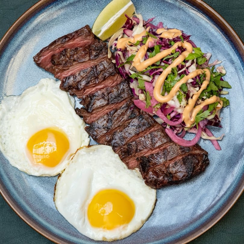 Sliced steak, two fried eggs, and a colorful salad with dressing and a lime wedge on a blue plate.